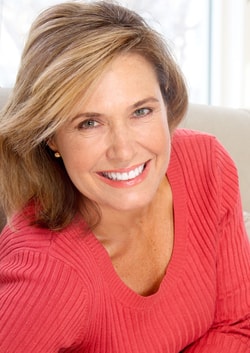 A woman with side swept brown hair and a coral sweater smiles warmly toward the camera