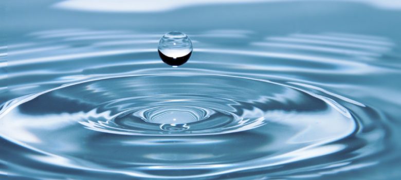 A drop of water is about to fall into clear blue water already rippling outward