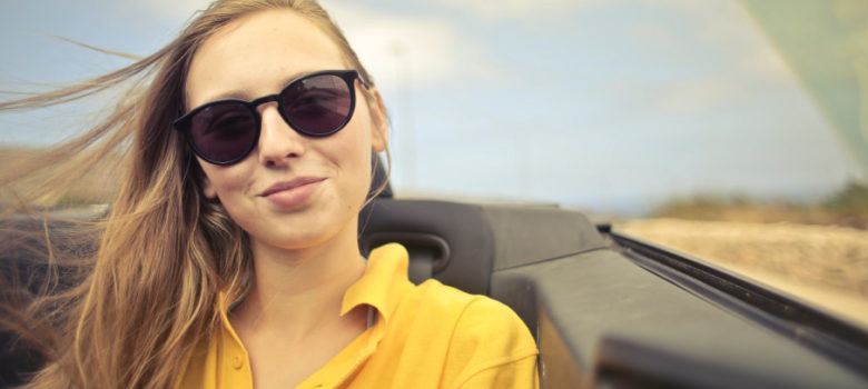 Blonde woman wearing sunglasses and a yellow blouse while sitting in the back seat of a convertible