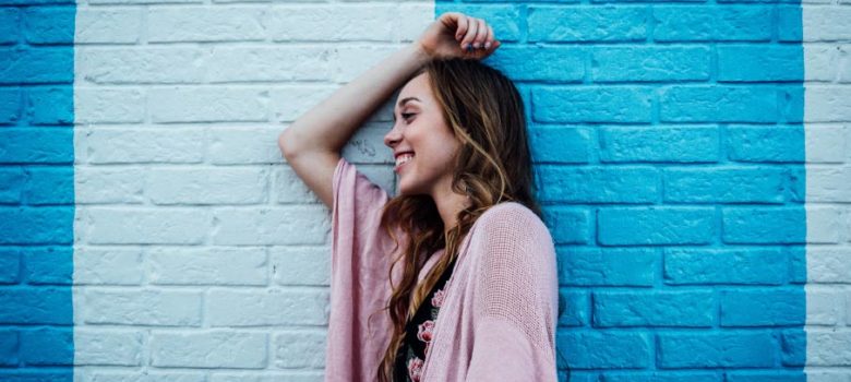 Brunette woman wearing a pink cardigan smiles as she leans against a brick wall painted 2 colors of blue